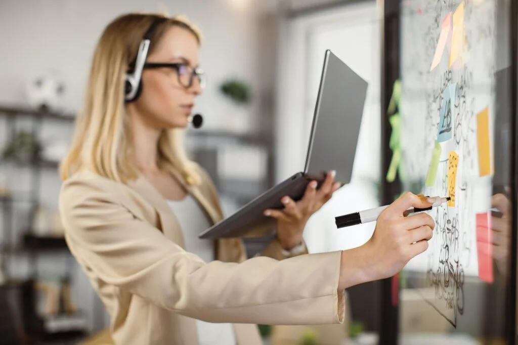 Woman in headphones with laptop writing on glass board