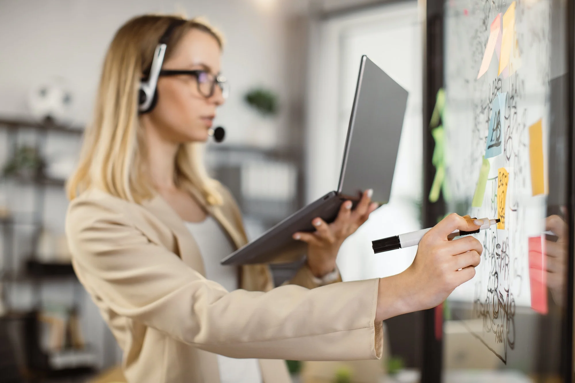 Woman in headphones with laptop writing on glass board